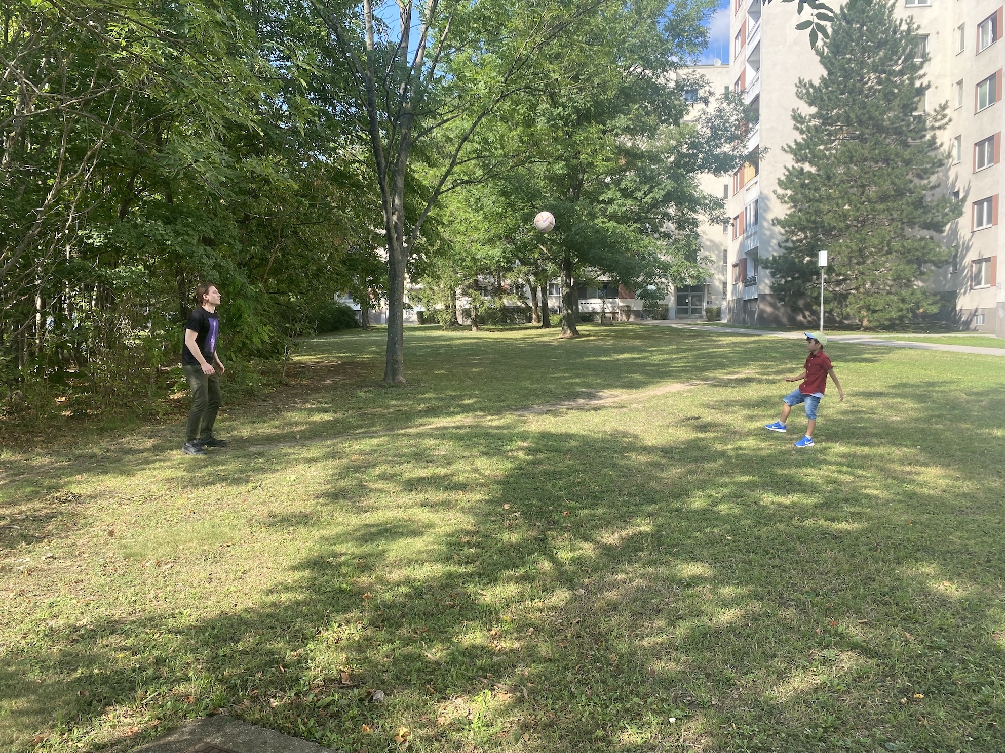 Auf einer Wiese spielen ein Kind mit einem Mann Fußball. Es ist sommerlich, Licht und Schatten liegen auf der Wiese, der Ball schwebt in der Luft, im Hintergrund eine Wohnsiedlung.