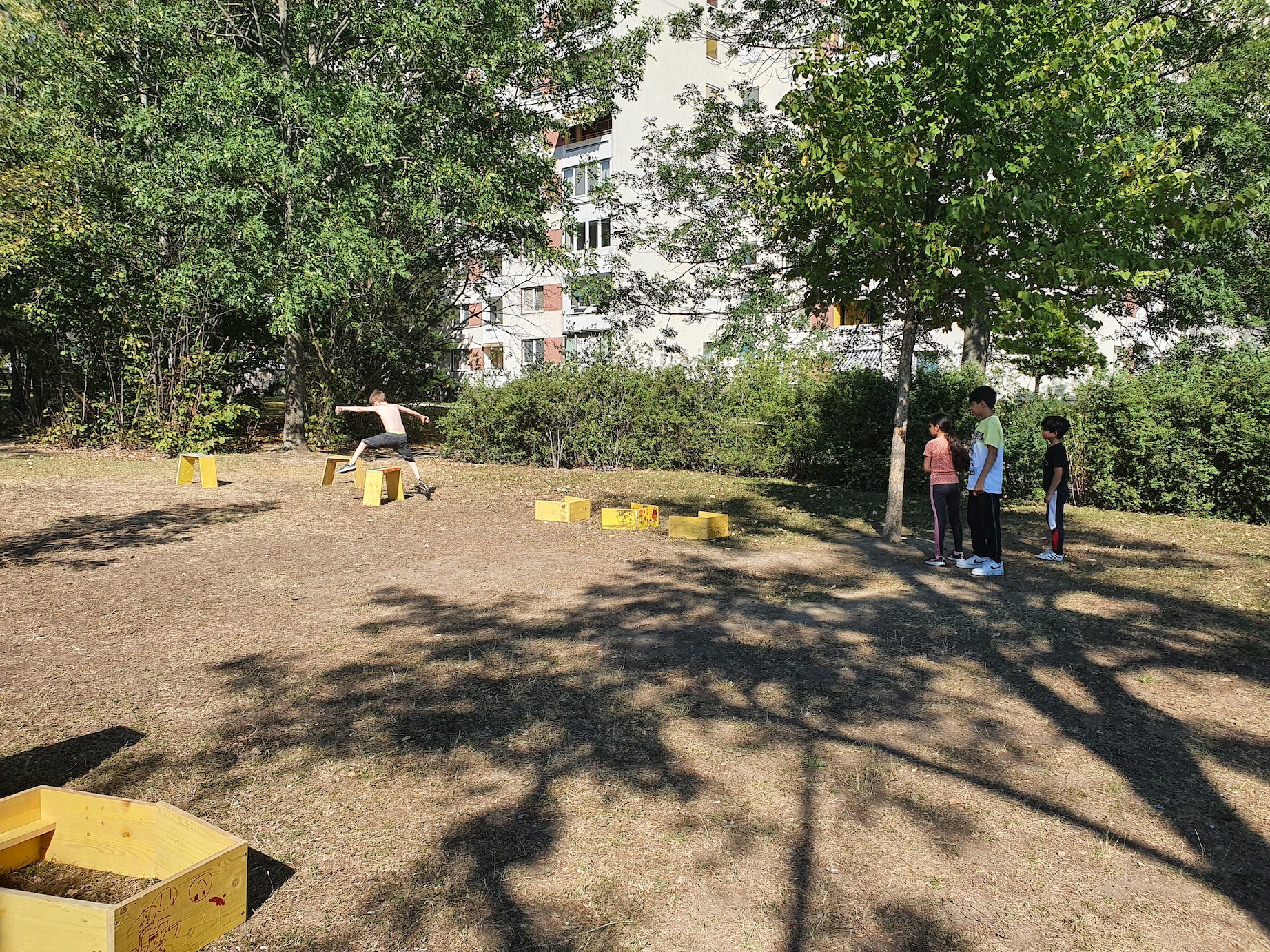 Kinder spielen im Hochsommer auf einer Wiese. Sie stehen in einer Schlange und warten darauf als nächstes durch den Parkour laufen zu dürfen. Die Wiese ist verbrannt, die Hitze des Tages dadurch spürbar.
