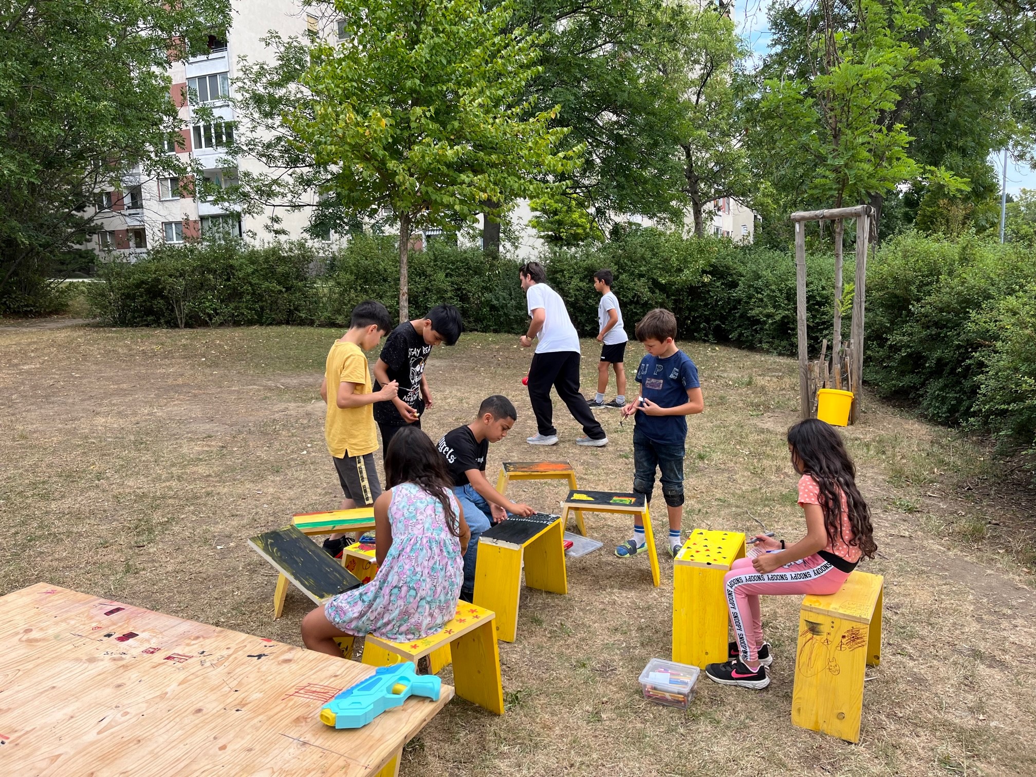 Kinder sitzen auf gelben Hockern auf einer Wiese. Dahinter wird ein Ballspiel gespielt. Es wird gemalt, geredet und geschaut.