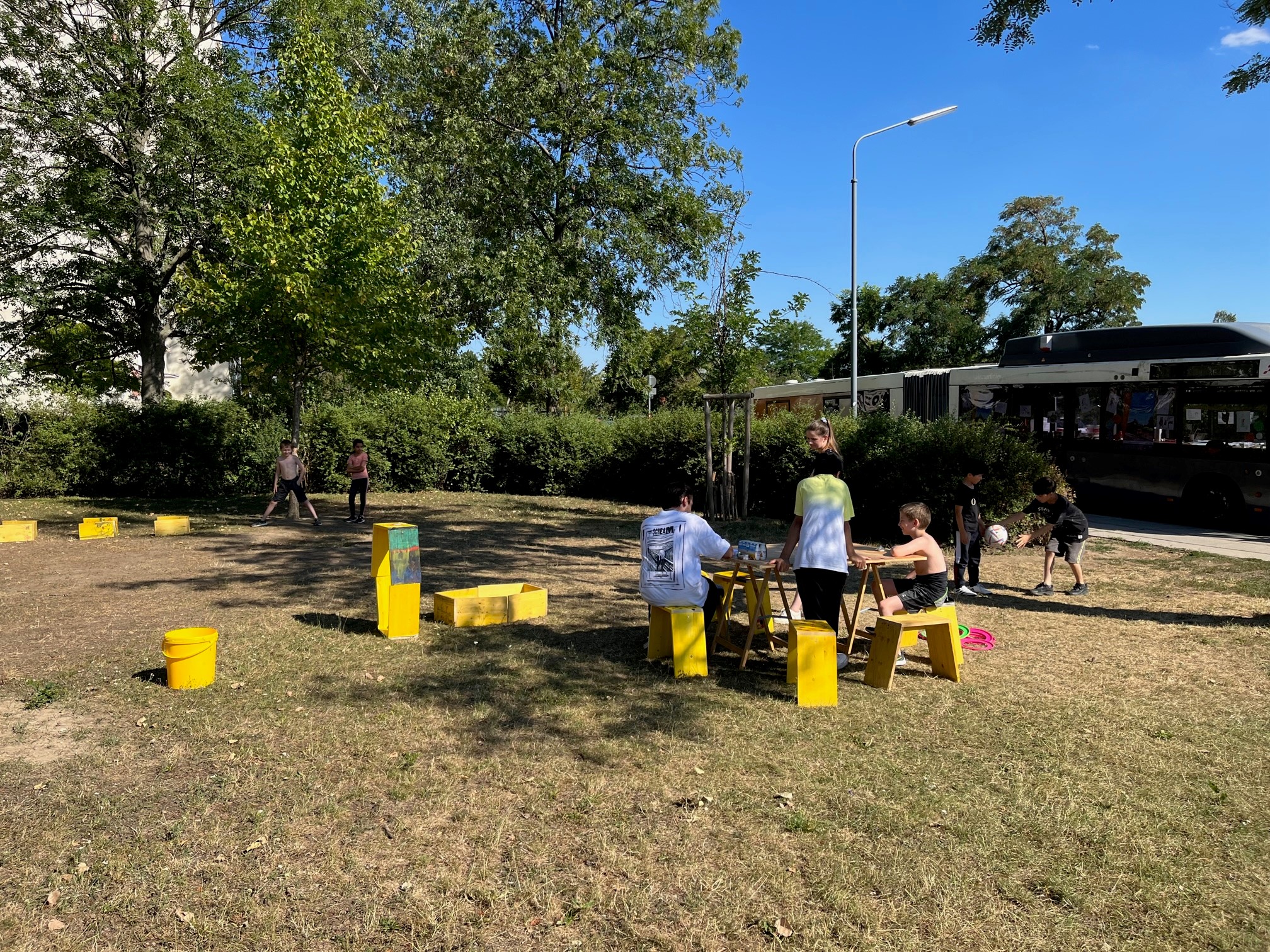 Kinder spielen auf einer Wiese wild in der Gegen, andere erholen sich auf Hockern sitzend und rasten.