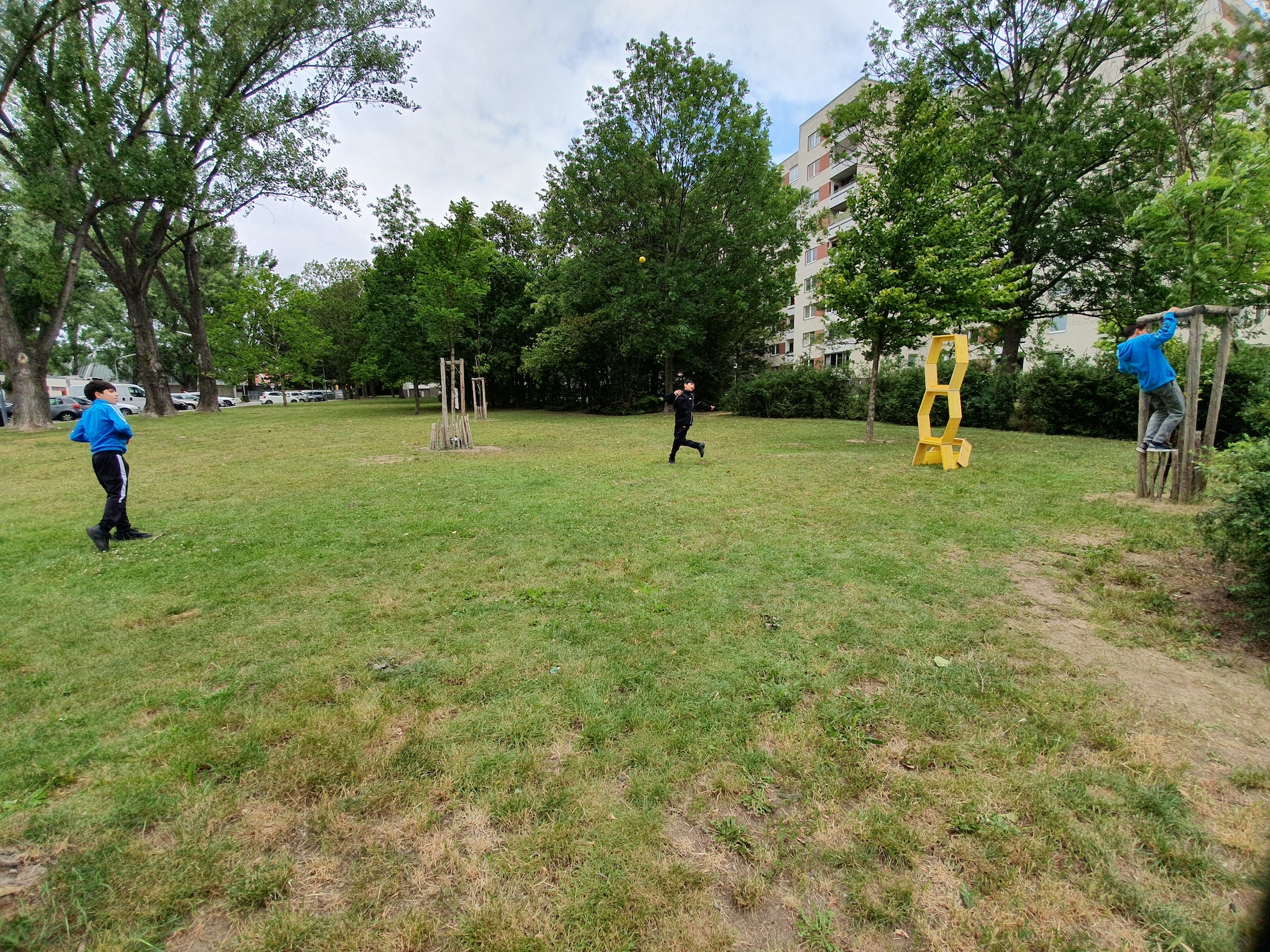 3 Kinder spielen auf einer Wiese. Eines hängt an einem Baum, ein zweites läuft, das dritte schaut zu. Eine Skulptur aus gelben Hockern ist gestapelt und dient als Zielscheibe.