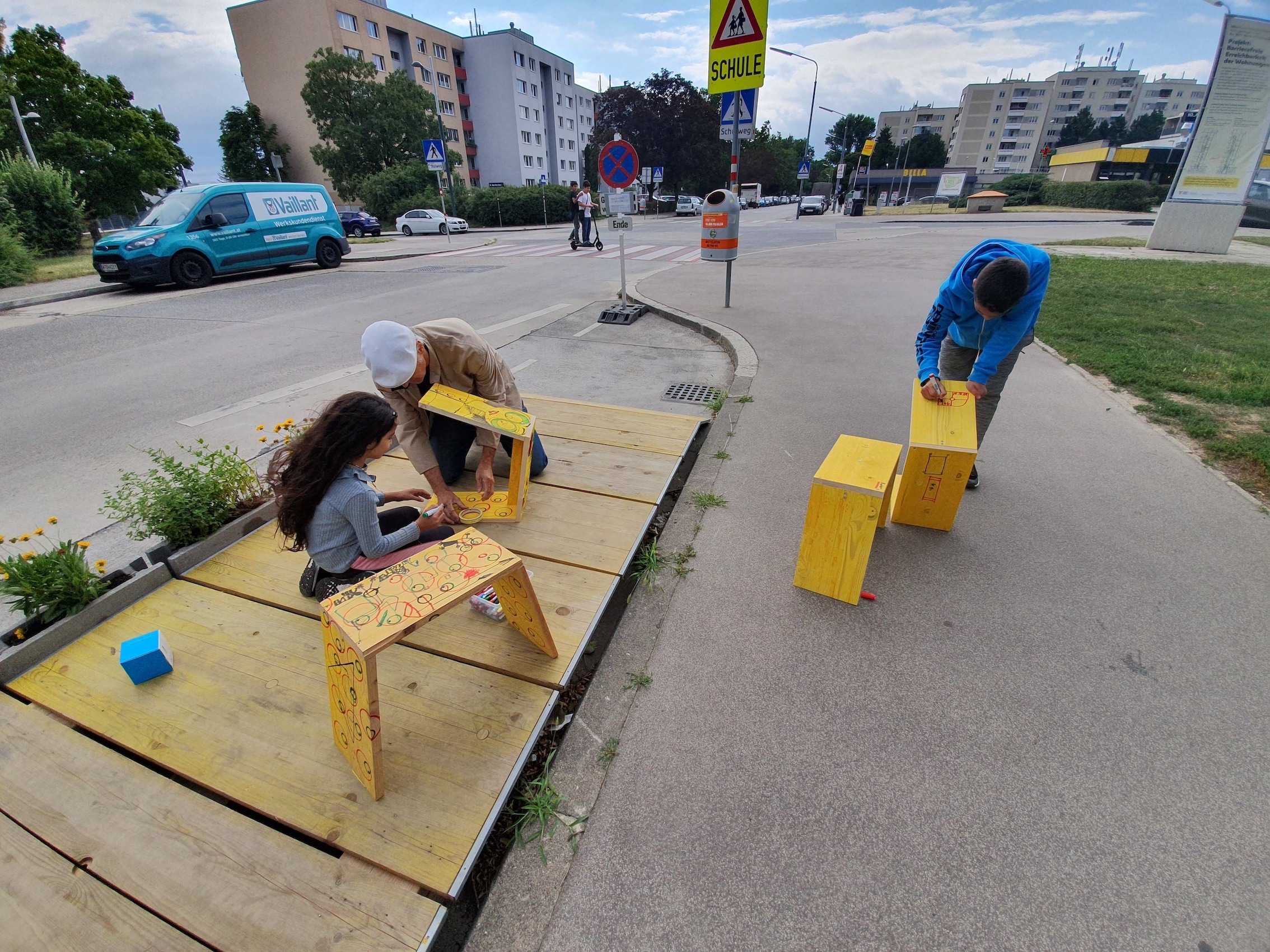 2 Kinder bauen und malen mit einem Erwachsenen gelbe Hocker an und sitzen dabei auf einer gelben Holzterrasse.