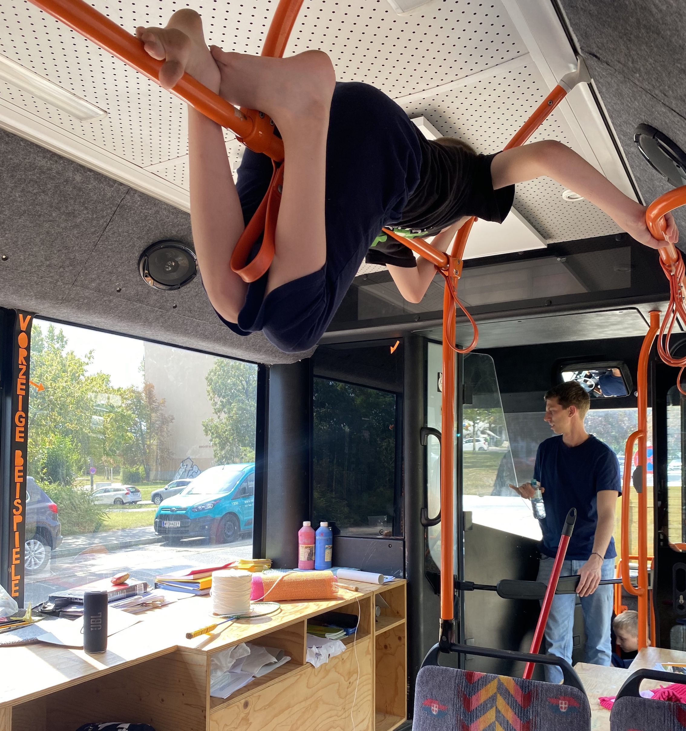 Foto aus dem Bus-Labor. Ein erwachsener Betreuer steht beim Fahrerbereich, unterhalb von ihm versteckt sich ein Junge, oberhalb von ihm hängt ein anderer Junge auf den eigentlich zum Anhalten gedachten Stangen.