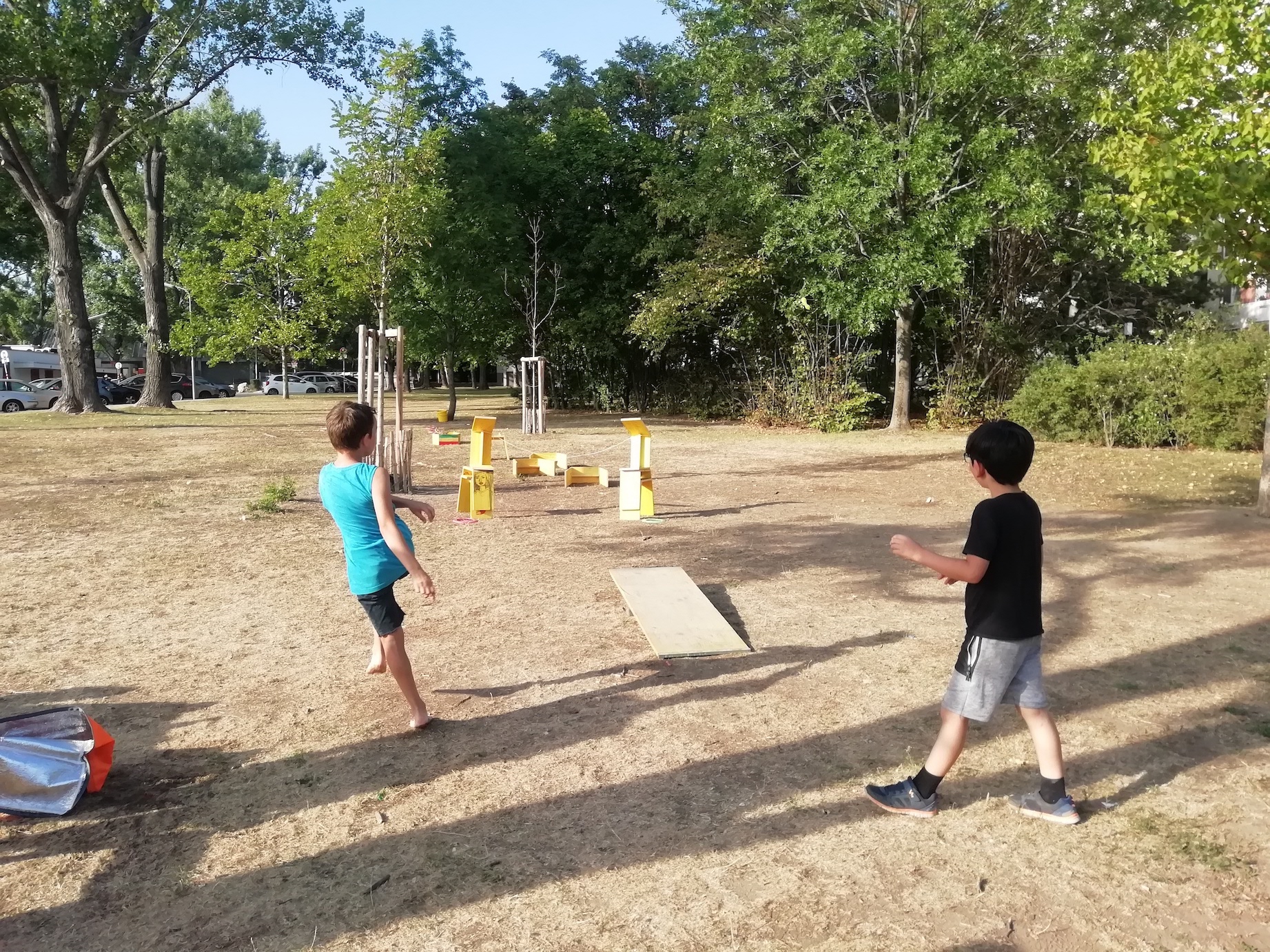 zwei Kinder stehen auf einer vertrockneten Wiese. Einer davon hat soeben gegeben einen Ball gekickt. Das Treffziel sind aus gelben Hockern gebaute skulpturale Elemente.