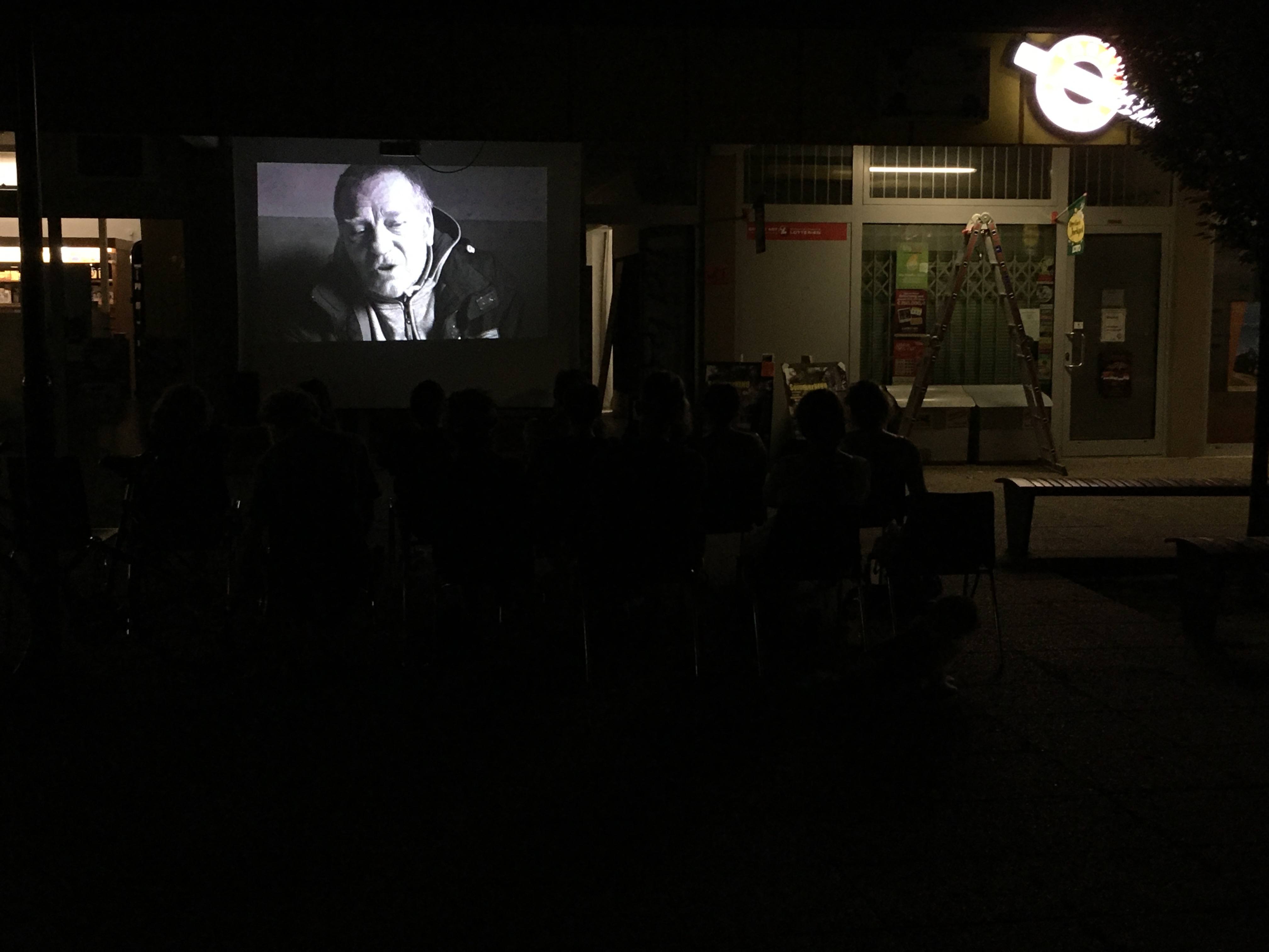 dunkles Foto einer Szene aus dem Film Aufzeichnungen aus der Unterwelt. Personen-Silhouetten sind sichtbar, sowie das Leuchten von Reklametafeln bei Nacht. Die Kinoleinwand hängt im Außenraum einer kleinen Einkaufspassage zwischen Apotheke und Trafik.